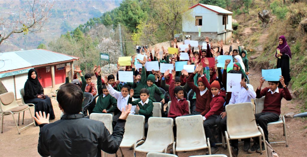 Students sitting in chairs outside and raising their hands to answer a question
