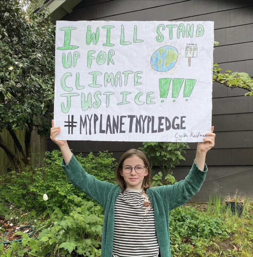 Person with black and white striped shirt and green sweater holding up a sign that says, "I will stand up for climate justice!!! #MyPlanetMyPledge"